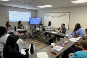 Image of college interns and CAPACares staff seated around a table in a conference room, with one person speaking.