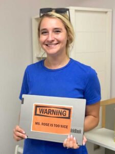Photo of a smiling blonde woman wearing a blue t-shirt, holding a laptop with an orange sticker that says "WARNING MS ROSE IS TOO NICE"