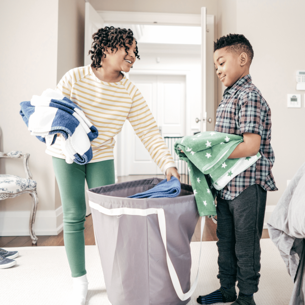 A young girl and a young boy are seen happily folding and sorting laundry together in a bright, well-lit room. The girl, wearing a yellow striped long-sleeve shirt and green leggings, is placing folded towels into a large gray laundry hamper. The boy, dressed in a plaid shirt and dark pants, is holding a stack of green towels with white stars. Both children are smiling, indicating they are enjoying the chore. The room is tidy, with a light-colored carpet and furniture in the background.