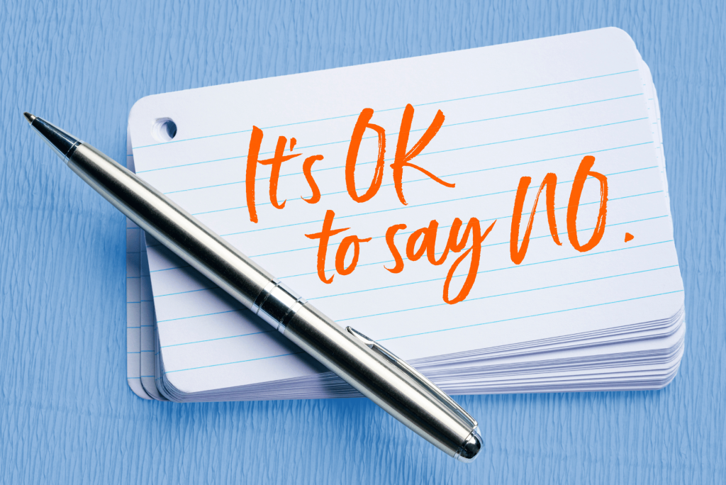 Image of a stack of index cards with "It's OK to say NO," written on them in large orange text. There's a silver ballpoint pen sitting diagonally across the corner of the cards, and they're sitting on a textured light blue background.