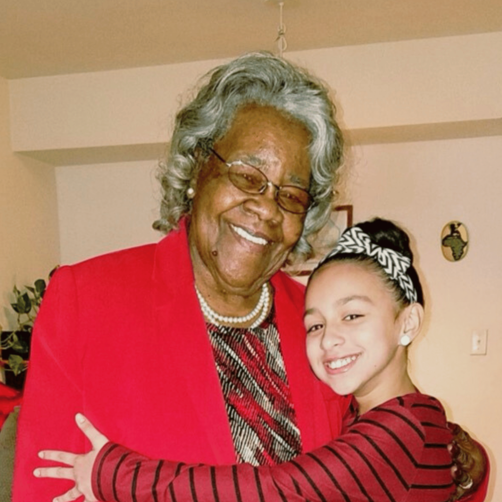 A photo of a smiling Black woman wearing a red blazer over a red, black, and white patterned blouse. She's wearing a pearl necklace, pearl earrings and glasses. She's being embraced by her preteen great-granddaughter, who is wearing a black and white striped headband and a red and black striped long-sleeved shirt.