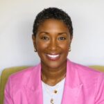 Professional headshot of a smiling Black woman wearing a pink blazer. She has short, dark, curly hair and is wearing gold jewelry.